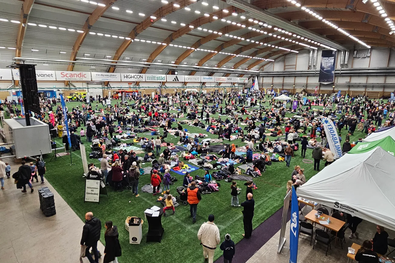 Massor av människor på en konstgräsyta inomhus, många sitter och har picknick.