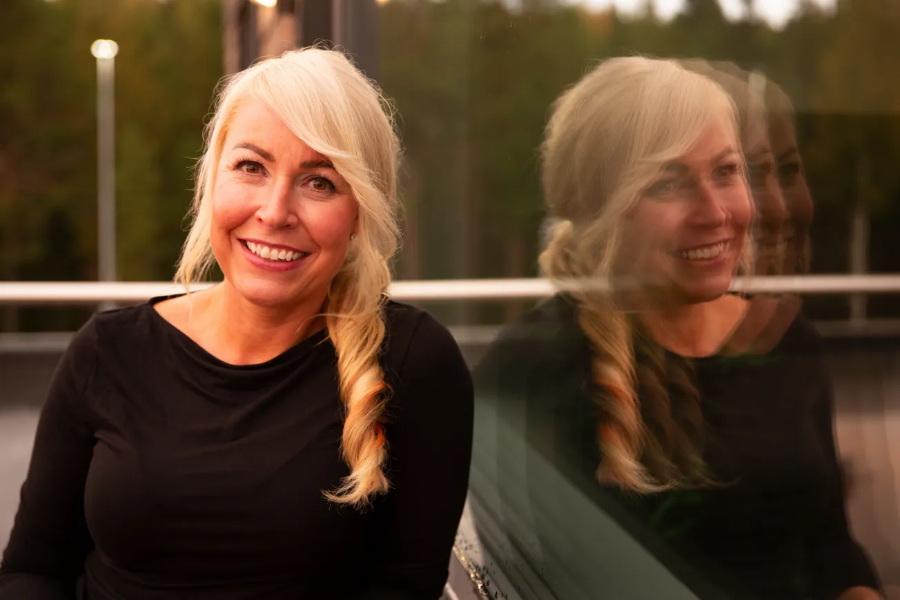 En kvinna ler in i kameran och sitter ner och lutar sig mot ett fönster.