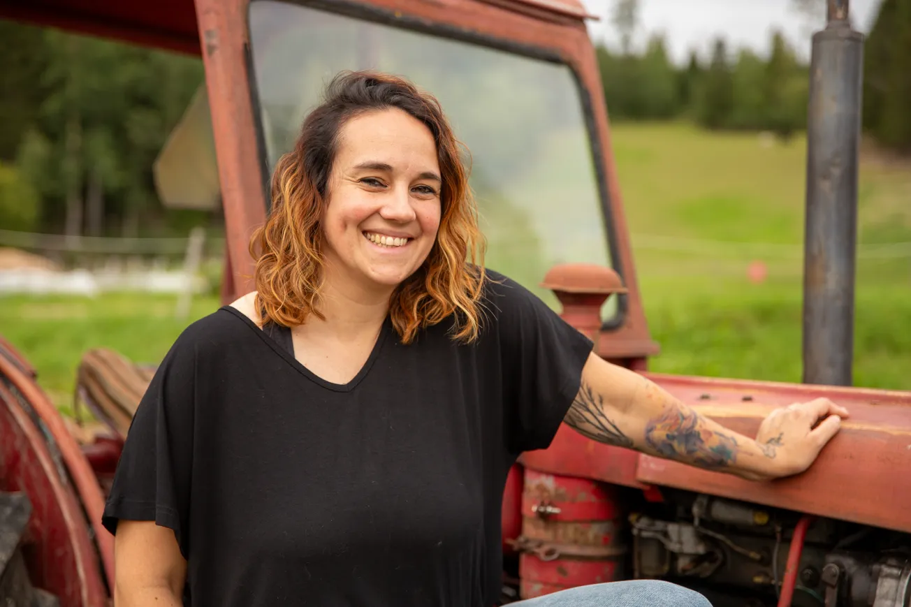 Porträttbild på kvinna som står bredvid en äldre traktor, tittandes in i kameran.