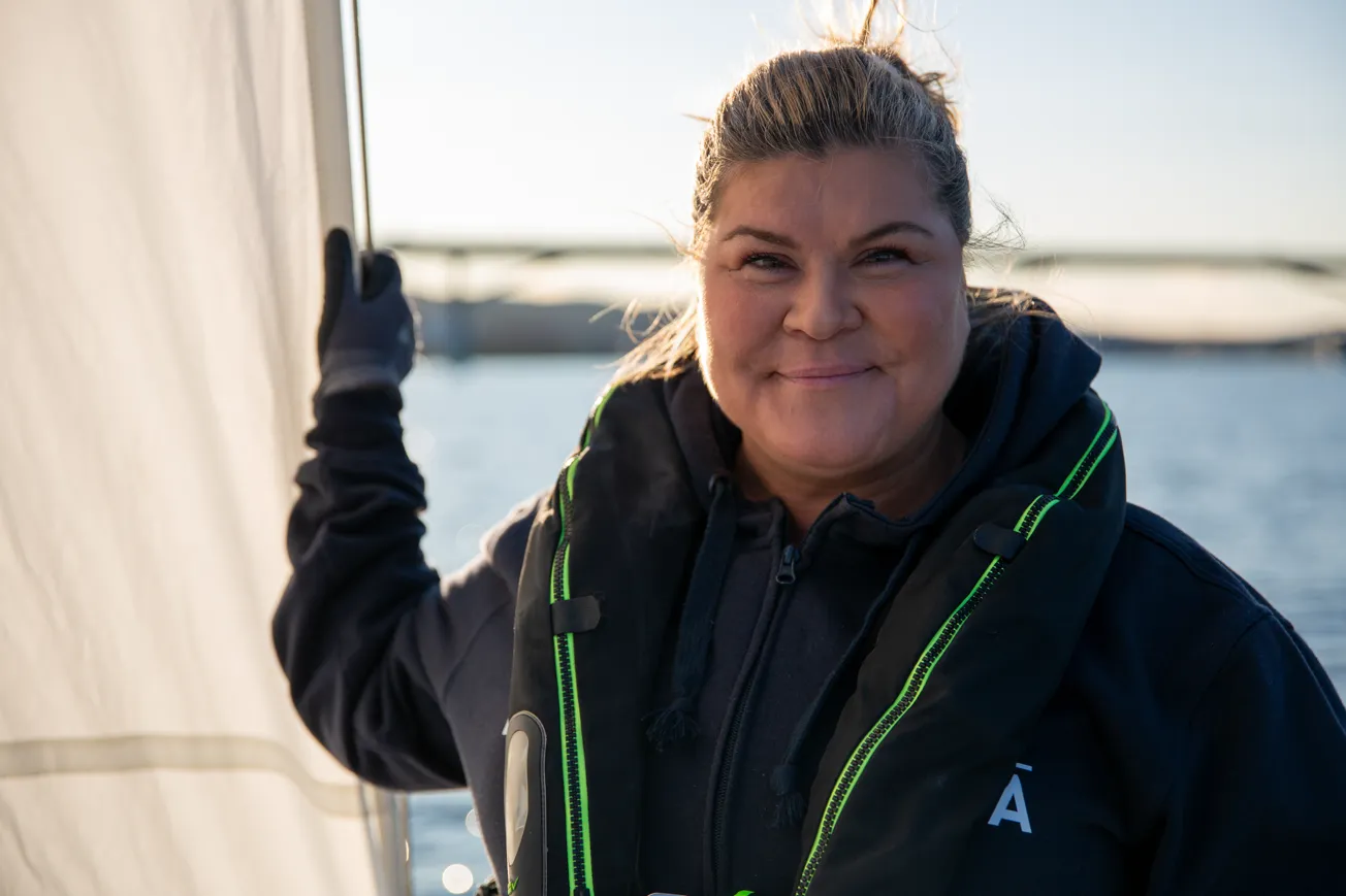 En kvinna tittar in i kameran och ler, medan hon står på en segelbåt och håller i sig i seglet. Man ser havet bakom henne.