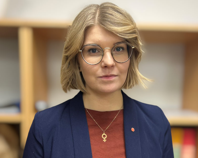 Kvinna med blå kavaj, röd tröja och glasögon. Hon har blont hår och ett halsband med kvinnosymbol.
