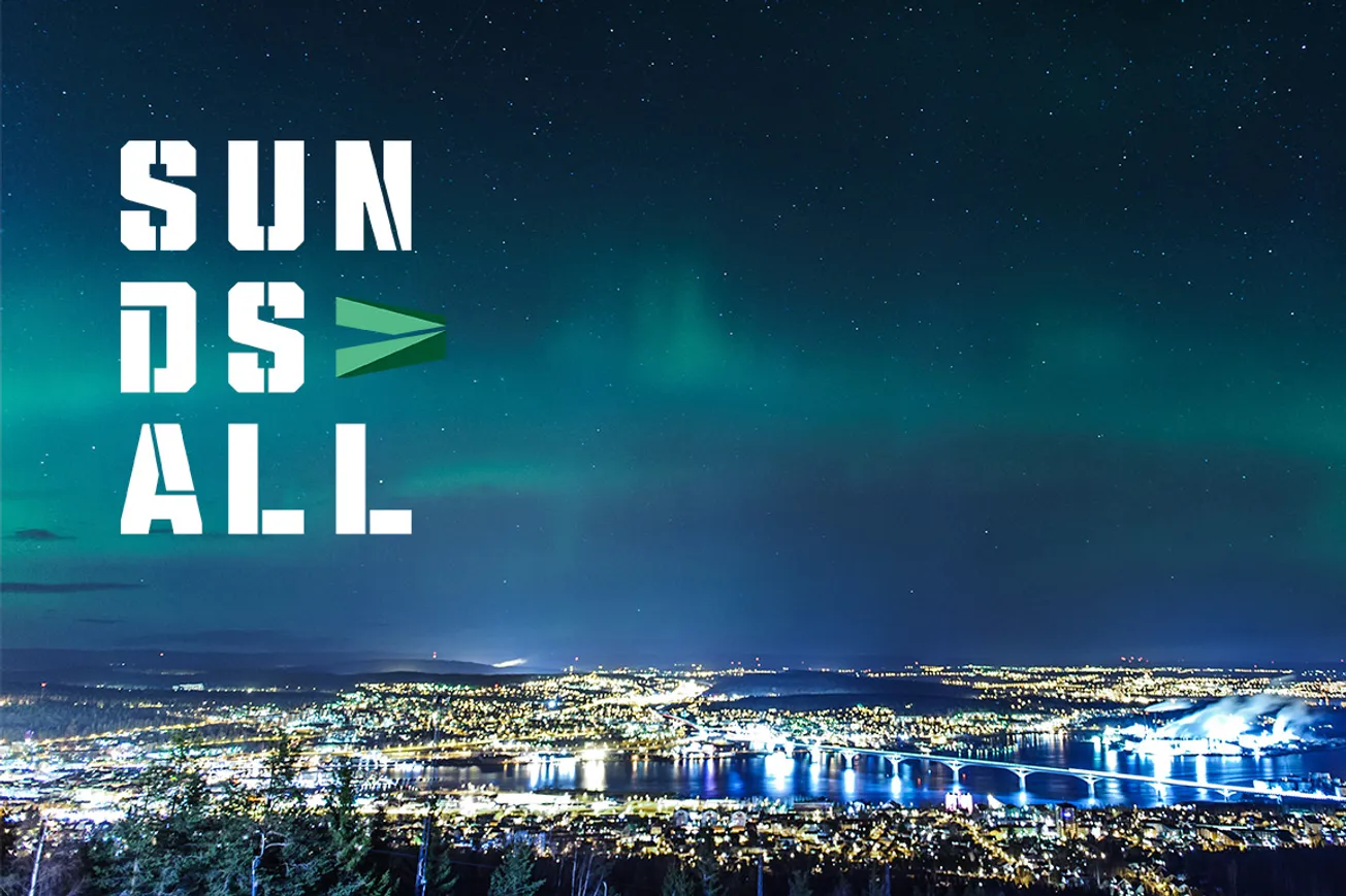 Nattvy över Sundsvall med upplyst stad, broar och vatten, fotograferad ovanifrån. Grönt norrsken syns över himlen och platsvarumärkets logotyp ”Sundsvall” är placerad till vänster i bilden.