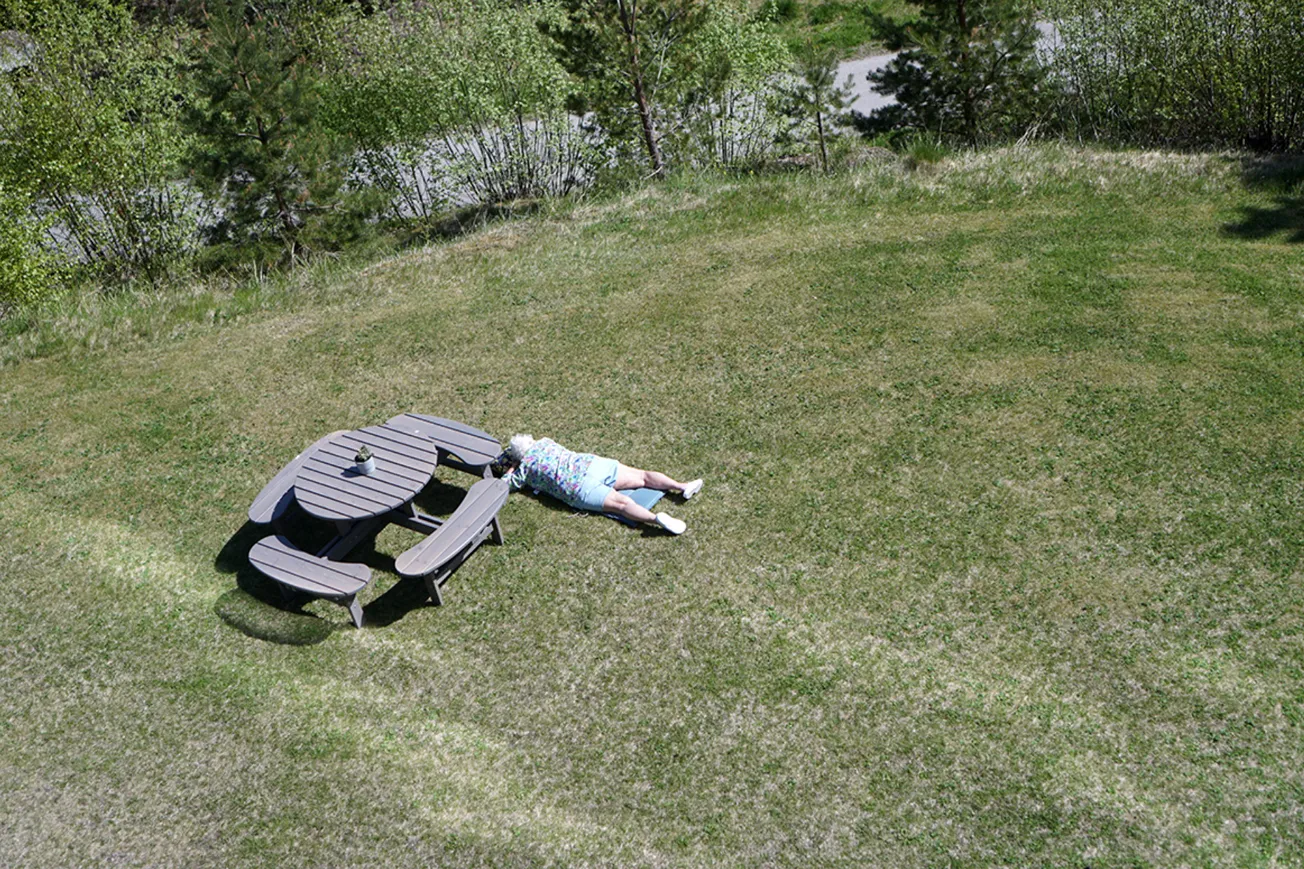Gräsmatta med ett runt bord och fyra bänkar. Till höger ligger en person på gräsmattan och solar.