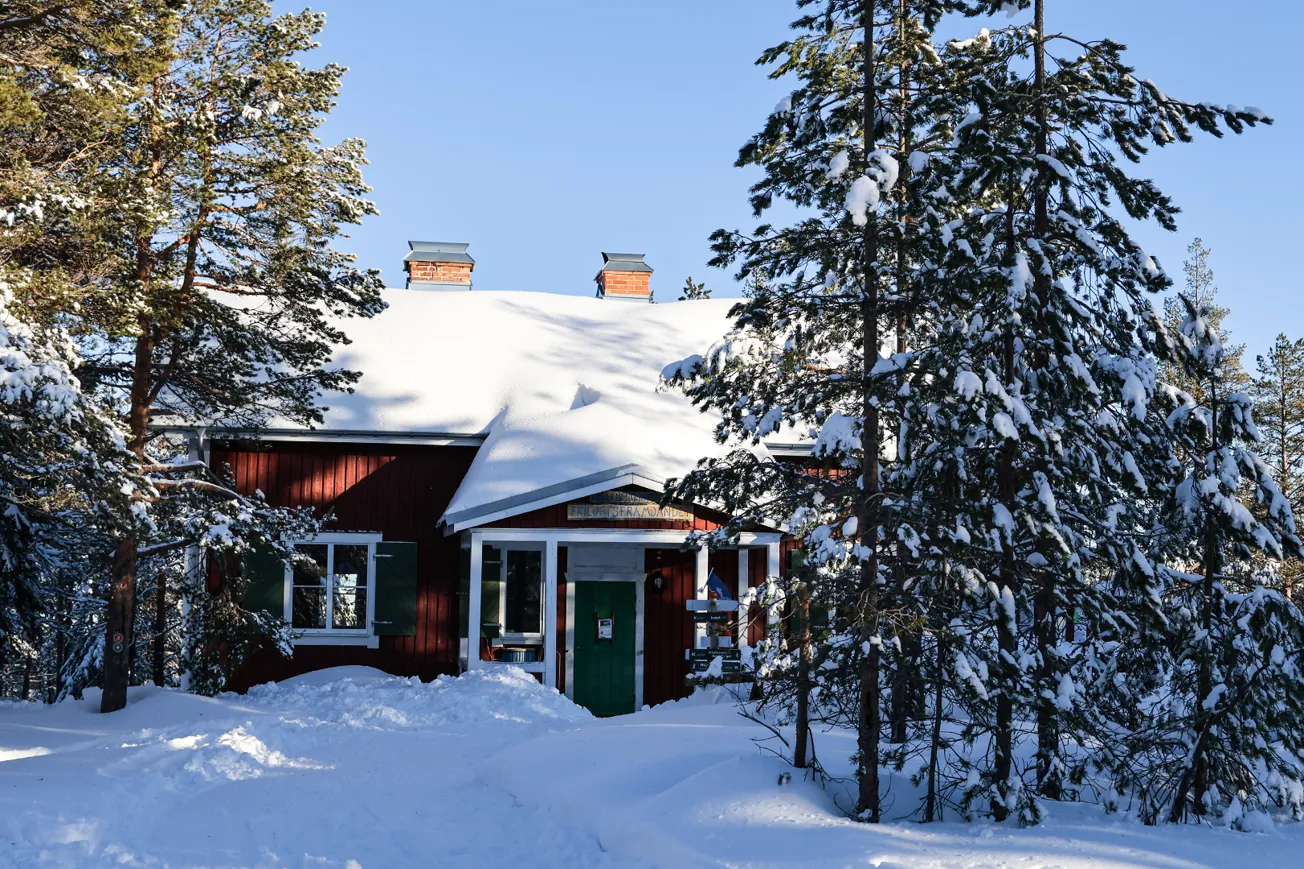 Röd stuga omgiven av träd i vintermiljö.