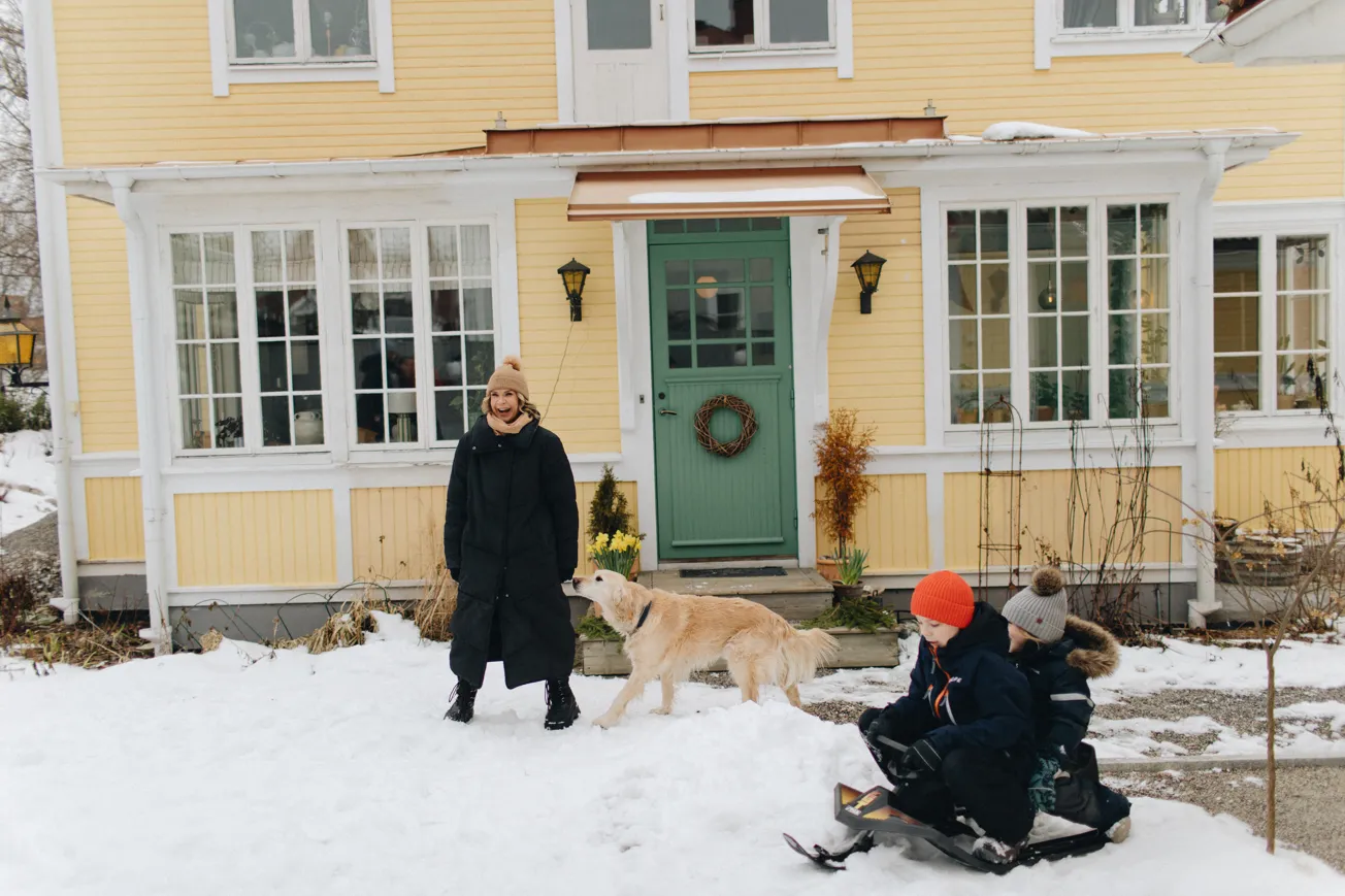 En mamma, en hund och två barn på snowracer leker ute i snön framför ett gult hus.