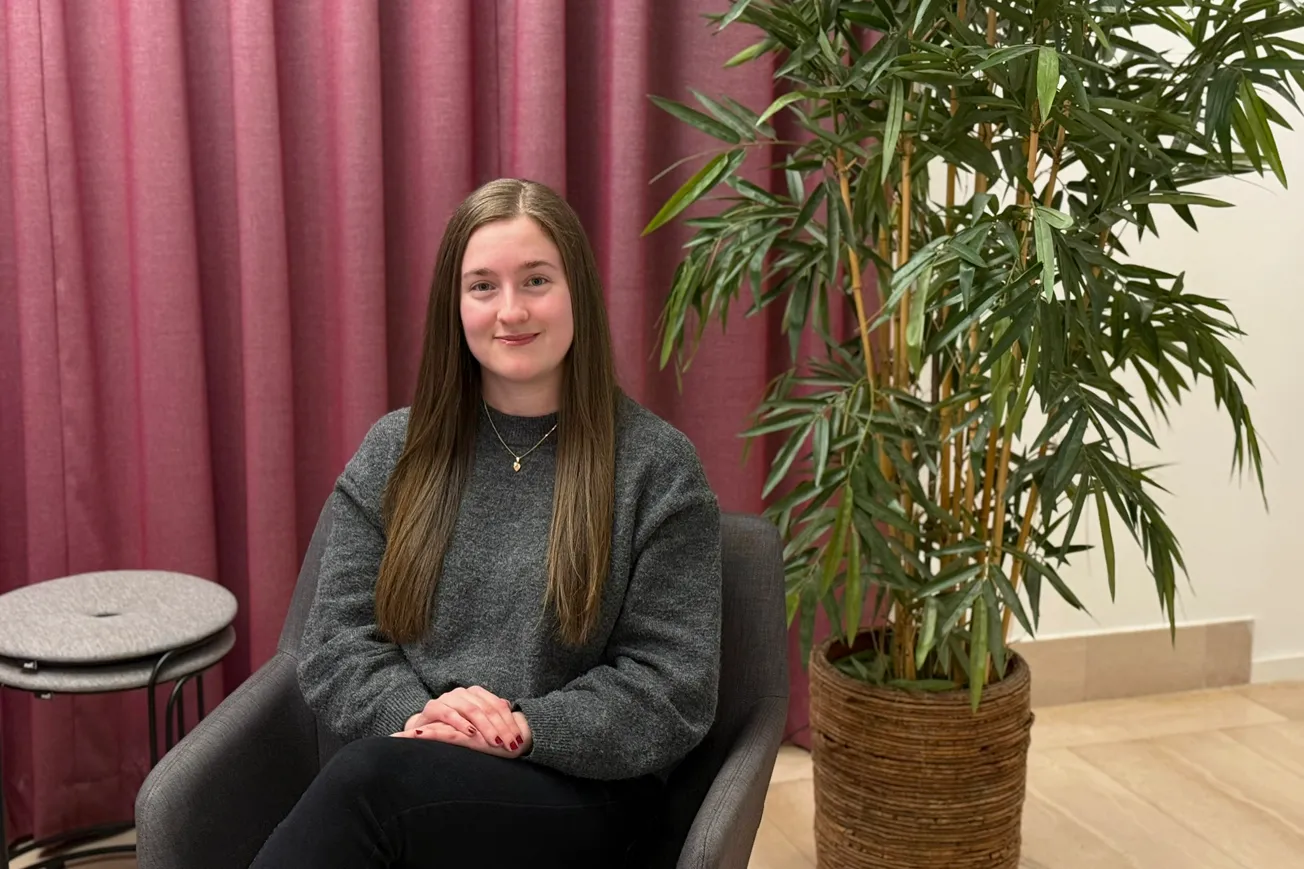 En tjej som tidigare studerat på  Yrkeshögskolan Mitt sitter i en grå soffa med lila gardiner i bakgrunden.