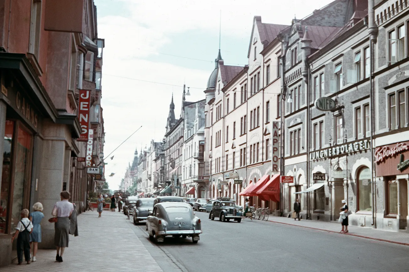 Gata med stenhus, parkerade bilar och folk som promenerar.