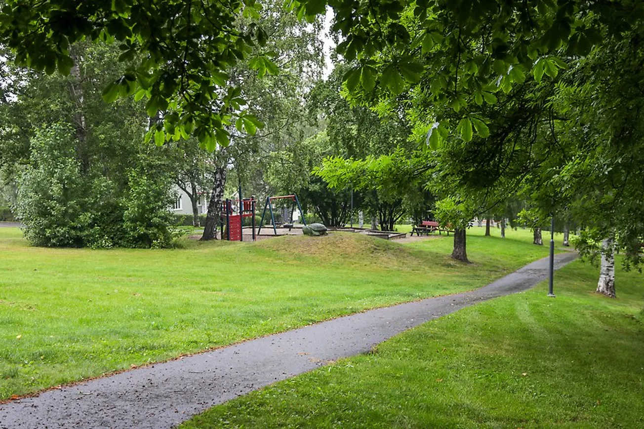 Bäckparken med grön gräsmatta, en stig och en lekmiljö i bakgrunden.