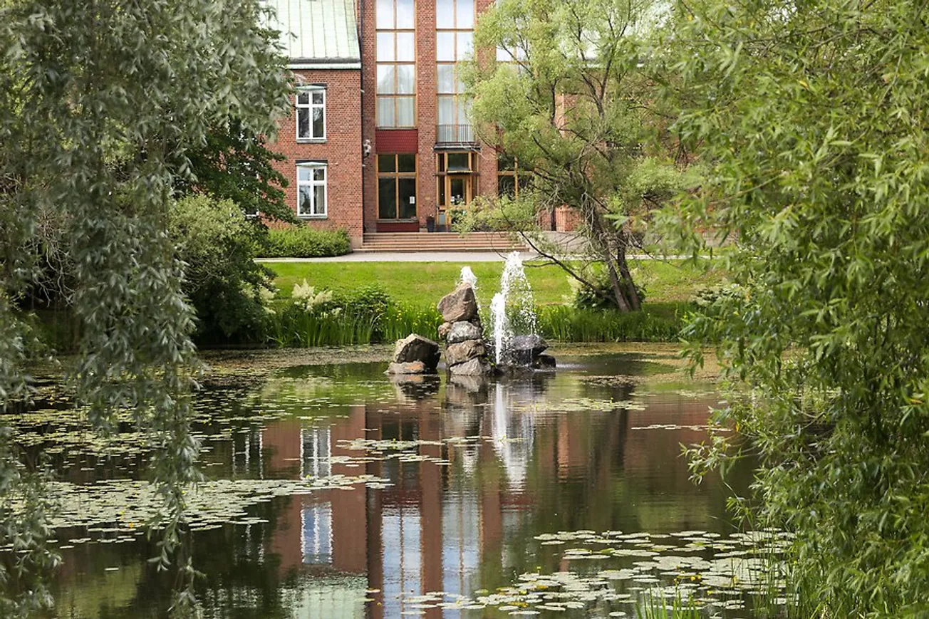 Bünsowska tjärnen sommartid där en damm syns i förgrunden framför en stor byggnad.