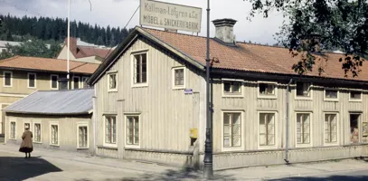 Trähus vid korsning. På taket sitter en stor skylt med texten Källman-Löfgren och Co Möbel och snickerifabrik