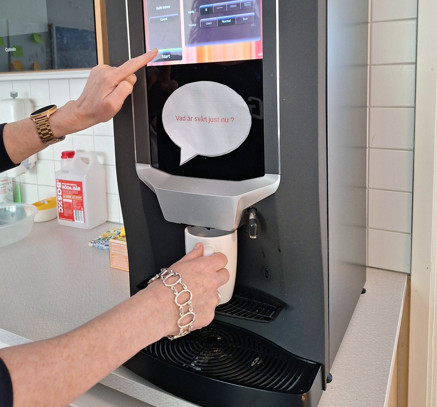 Närbild på händer vid en kaffemaskin där en kopp fylls samtidigt som en lapp på maskinen visar texten ”Vad är svårt just nu?”.