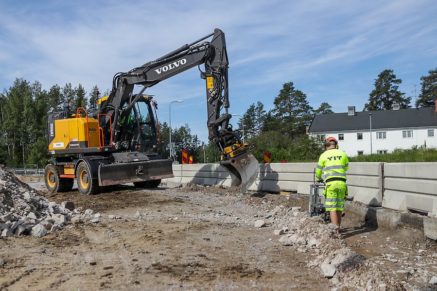 En gul och svart Volvo-hjulgrävare arbetar på en byggarbetsplats vid en väg under en klarblå himmel. En markarbetare i neongul varselklädsel och skyddshjälm står i förgrunden och använder en mindre maskin vid sidan av en rad betongbarriärer. I bakgrunden syns gröna träd och vita bostadshus.