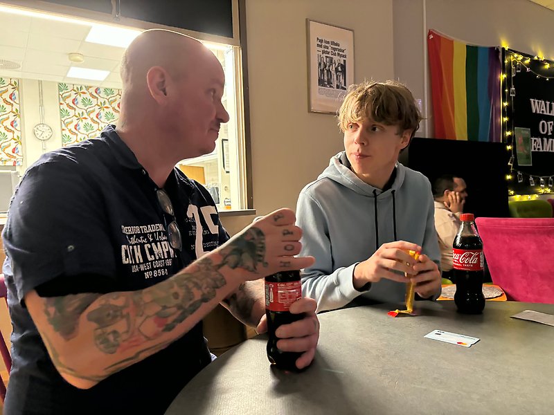 En man sitter vid ett bord med en yngre kille. Båda fikar.