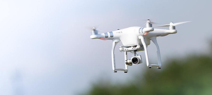 Fotografi som visar en vit drönare som flyger över ett skogslandskap, suddig bakgrund