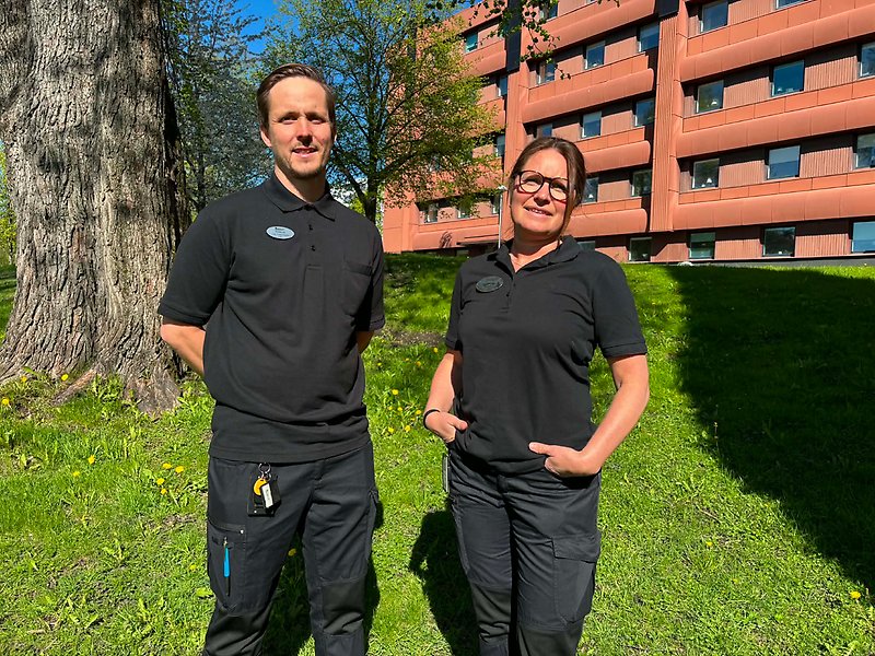 En man och en kvinna står bredvid varandra i en park framför ett rött hus.