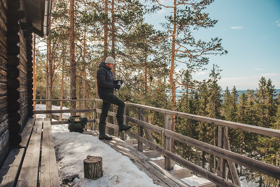 En man står på bron vid en friluftstuga och tittar ut över ett skogslikt vinterlandska.