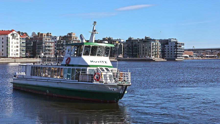 Foto som visar en elfärja med namnet Movitz som åker på vattnet i Sundsvalls inre hamn. I bakgrunden skymtar hus längs med kajen.