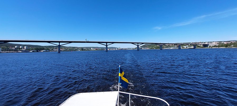 Fotot visar bakdelen på en båt med svensk flagga i aktern. Bakom båten skymtar vatten och Sundsvallsbron