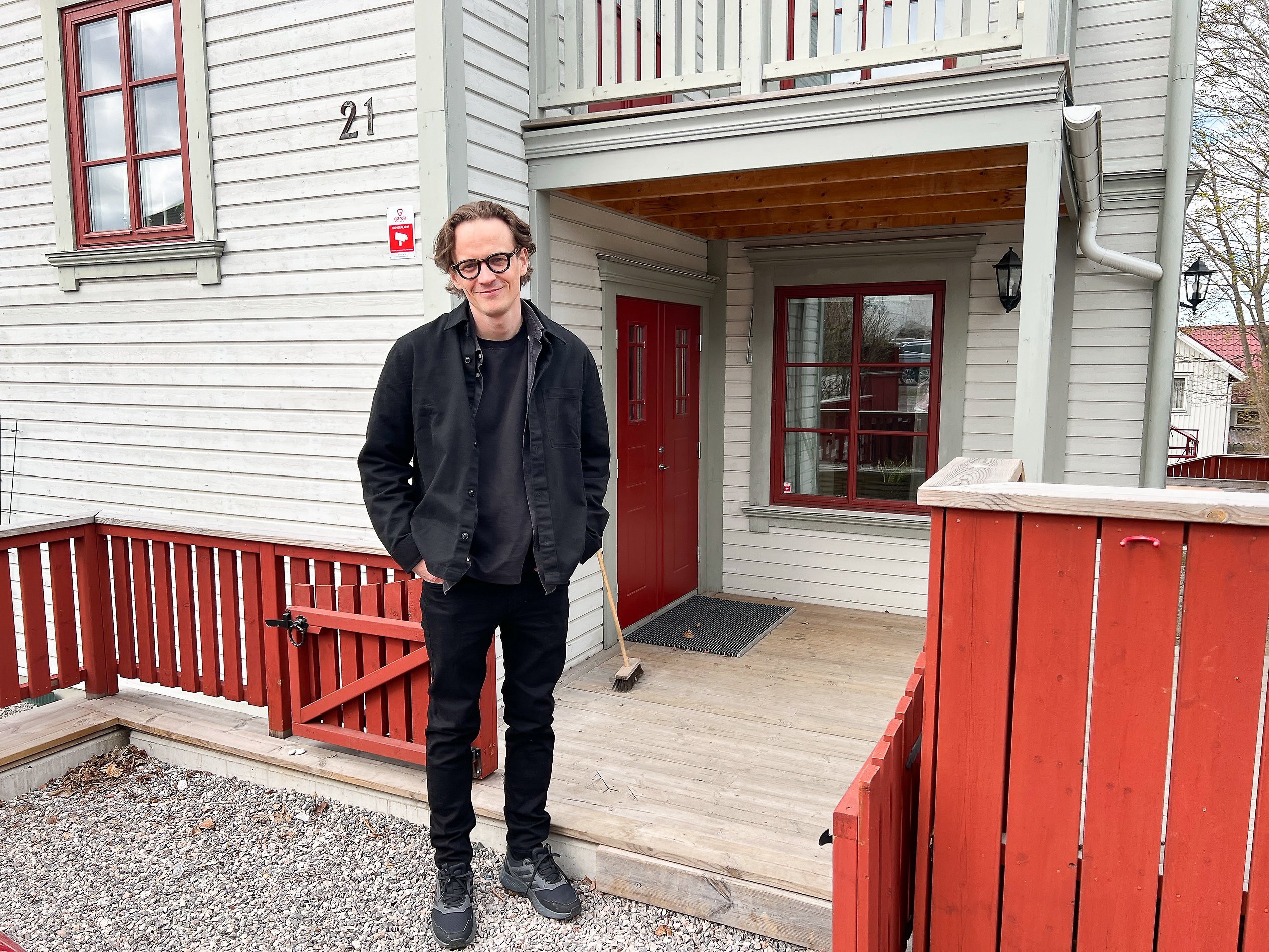 Man står med händerna i fickorna framför ett grått hus med röda knutar