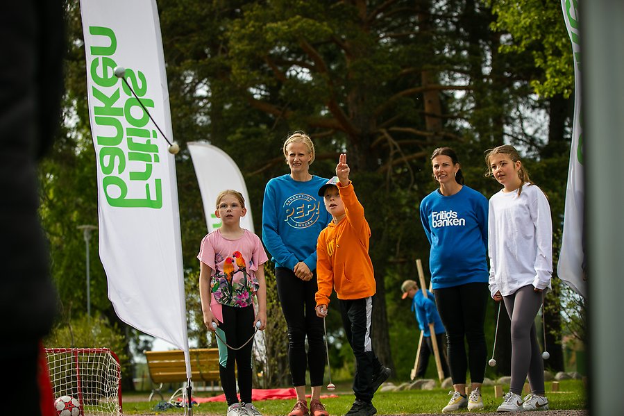 En grupp människor med vuxna och barn står på en gräsmatta. Runt om dem finns ett litet mål och en fotboll. De kastar en boll.