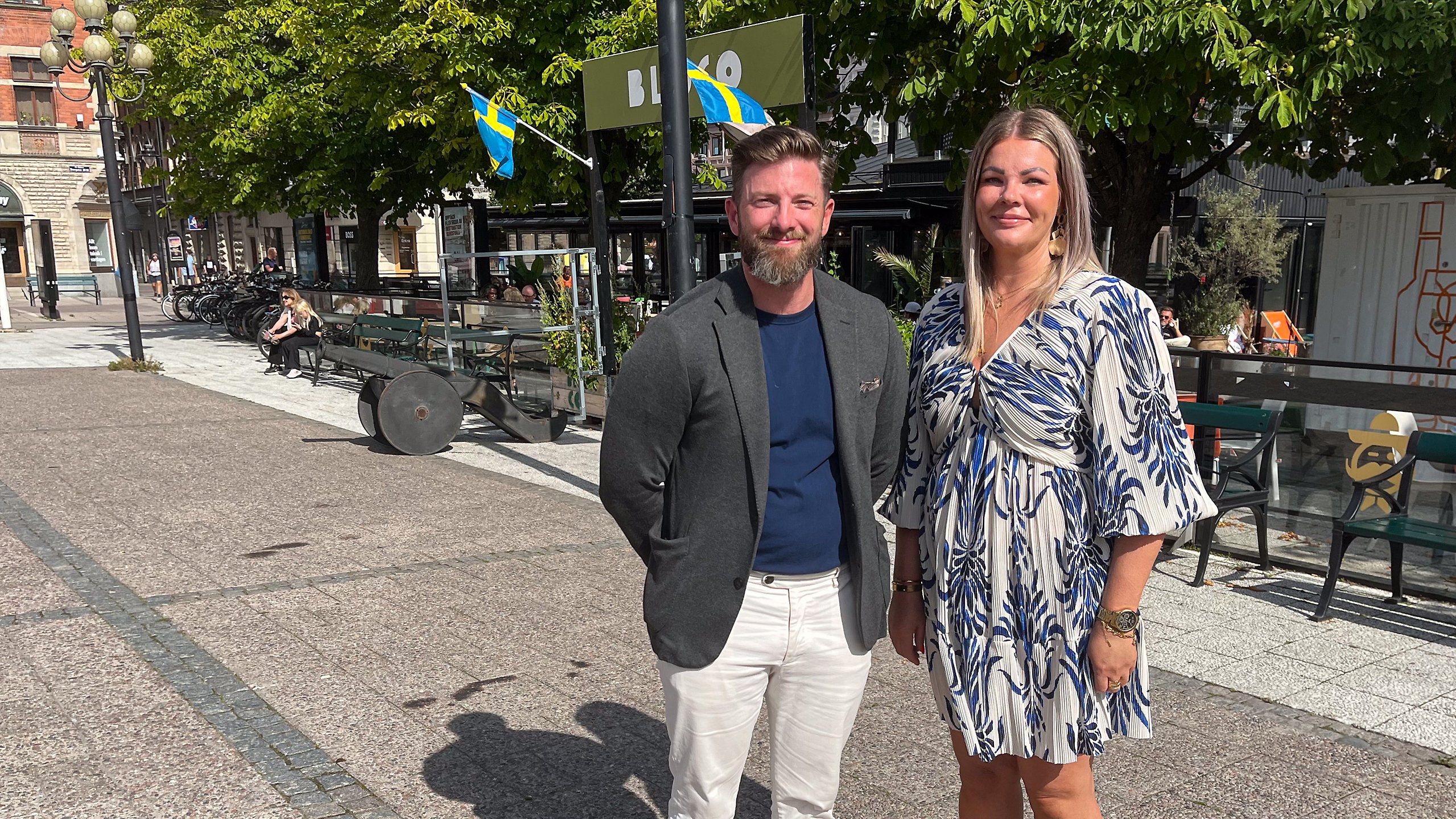 En man och en kvinna står på ett torg och kollar in i kameran.