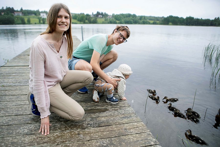 En kvinna och en man sitter på huk på en brygga tillsammans med ett litet barn. I vattnet nedanför bryggan simmar några änder.