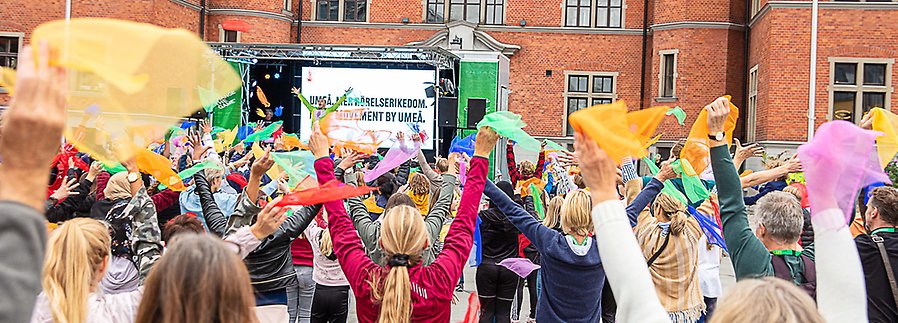 Foto som visar en folkmassa med ryggarna vänd mot kameran. I händerna på människorna på bilden håller de upp skira, färgglada tygbitar i luften. I bakgrunden syns en scen. 
