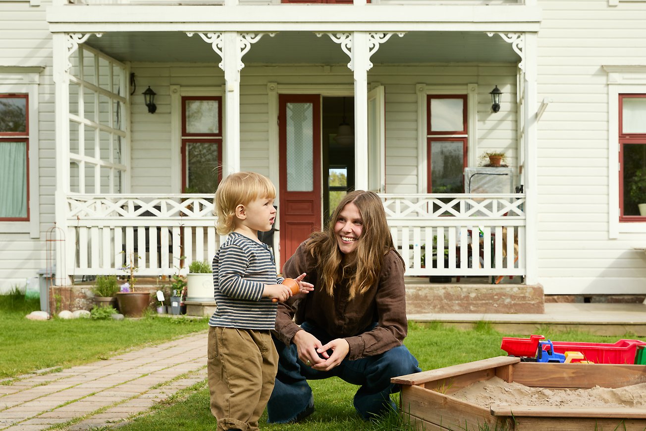 En mamma sitter på huk bredvid sitt lilla barn framför ett vitt hus.