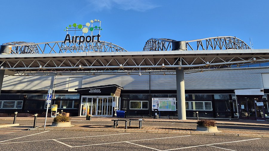 Foto som visar Sundvall-Timrå airports entré med fasadskylt och blå himmel. 